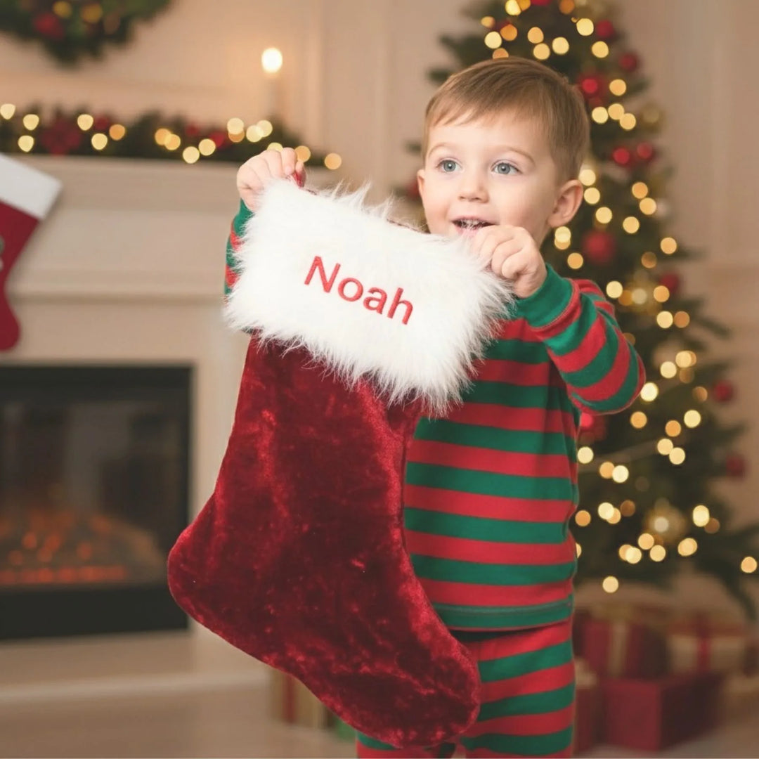 Child in striped pajamas holding a red Christmas stocking with 'Noah' on it, in front of a decorated fireplace and tree.