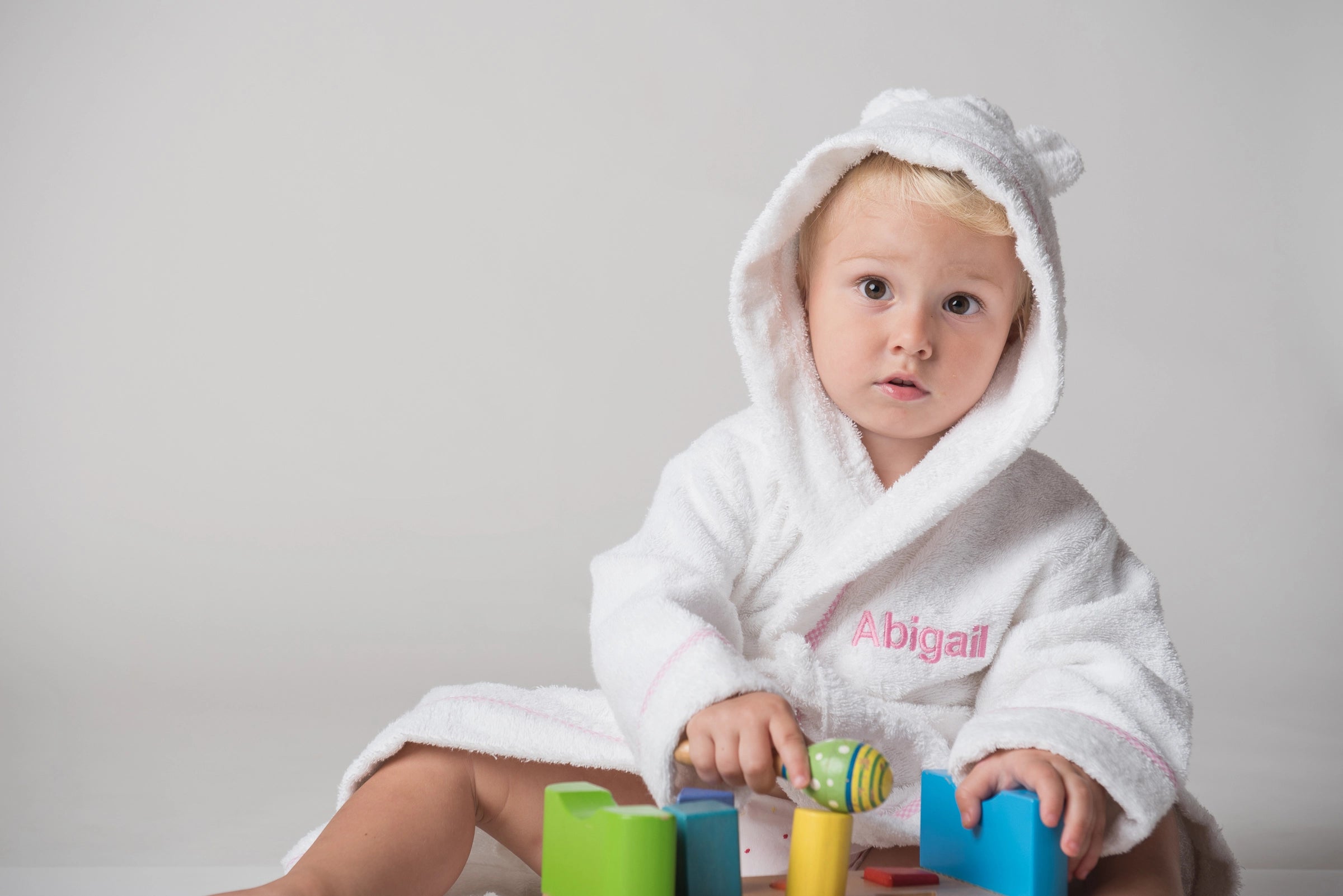 	Adorable baby girl smiles, wearing a soft white robe with delicate pink embroidery, playing joyfully with bright toys.