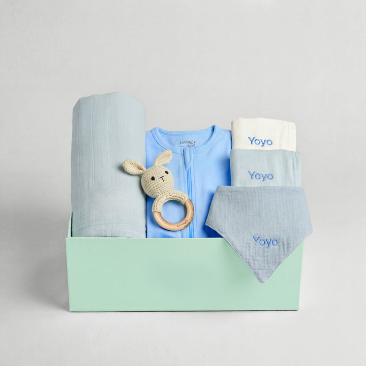 Baby gift set with blue outfit, blanket, and rattle in a box on a light gray background