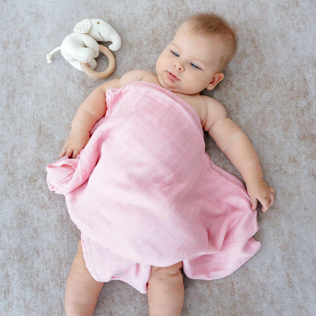 Baby covering in pink bamboo muslin swaddle and playing with Elephant Rattle Toy