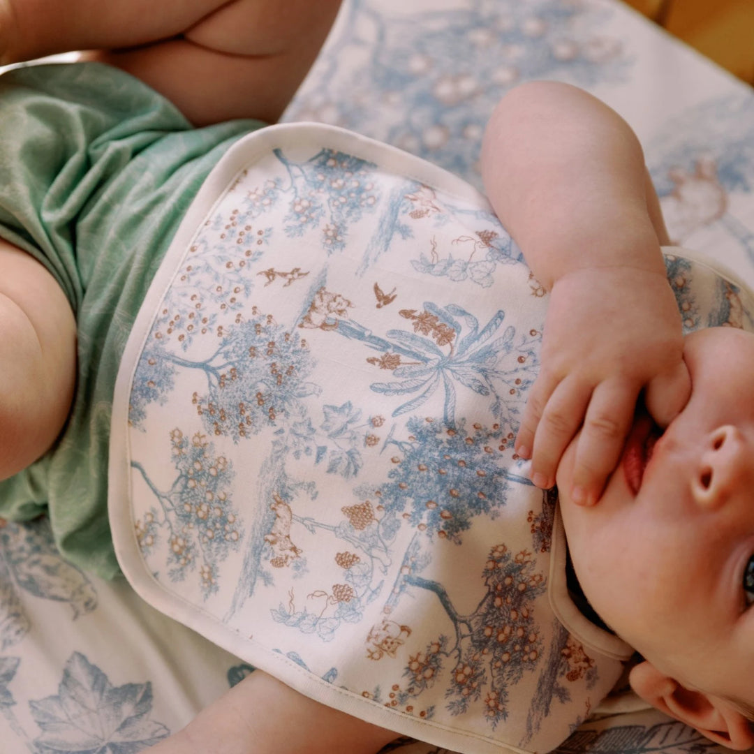 Baby wearing the House of Holly Fruit Toile reversible bamboo drool bib in blue, shown in a soft lifestyle setting.