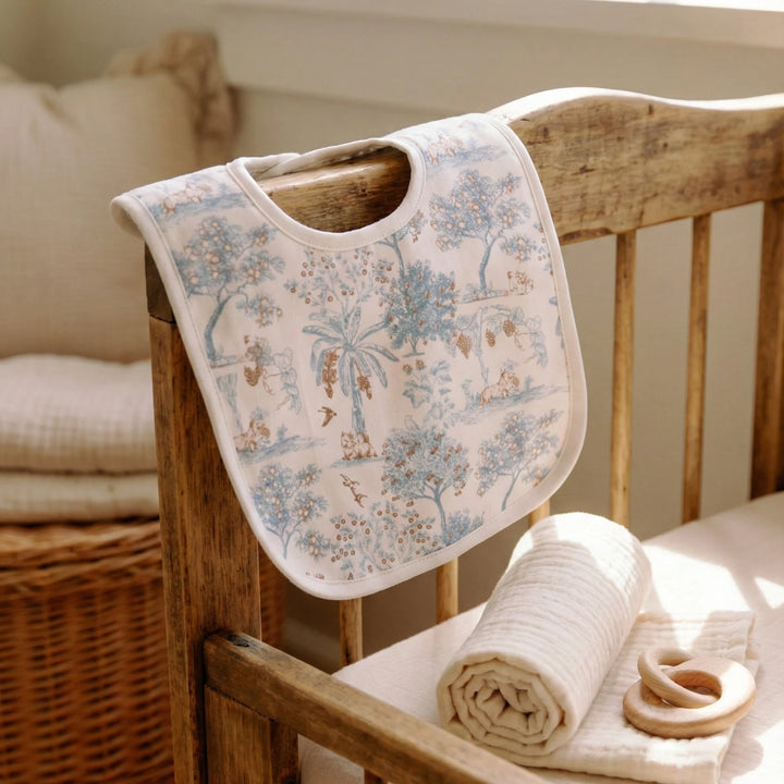 House of Holly Fruit Toile reversible bamboo drool bib in blue displayed over a wooden cot in a neutral nursery setting.