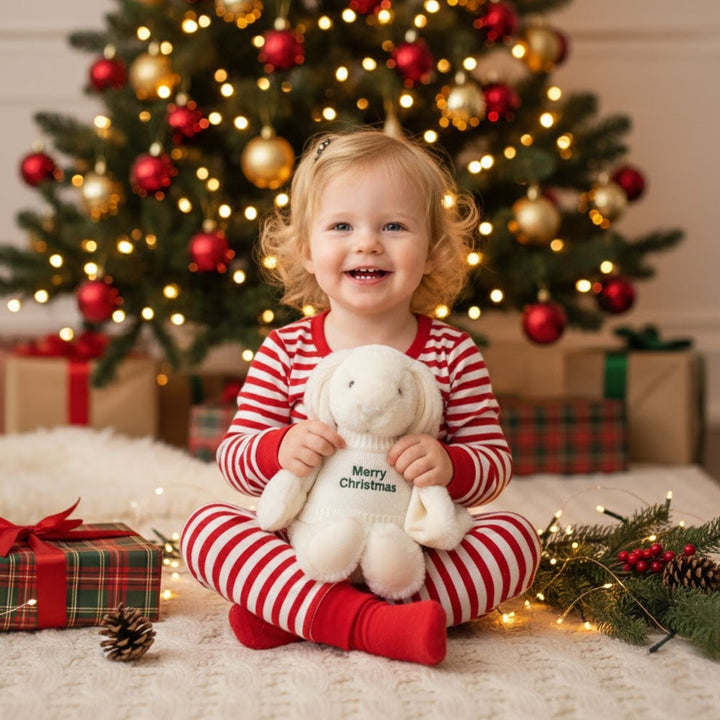 Child in red and white striped pajamas holding a plush toy in front of a decorated Christmas tree.