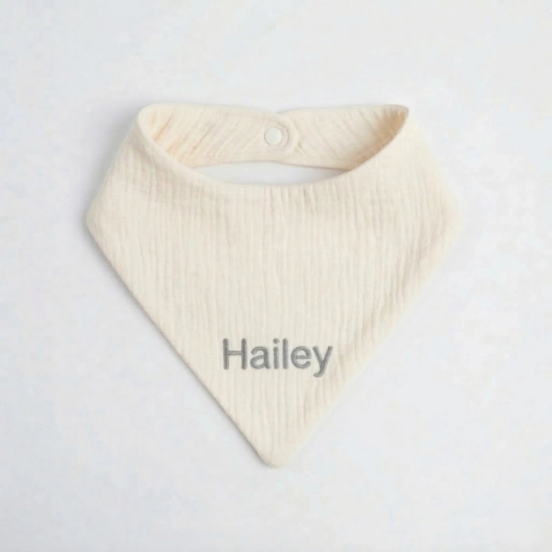 Personalised cream muslin baby bib embroidered with the name “Hailey” in grey thread, photographed on a clean white background.