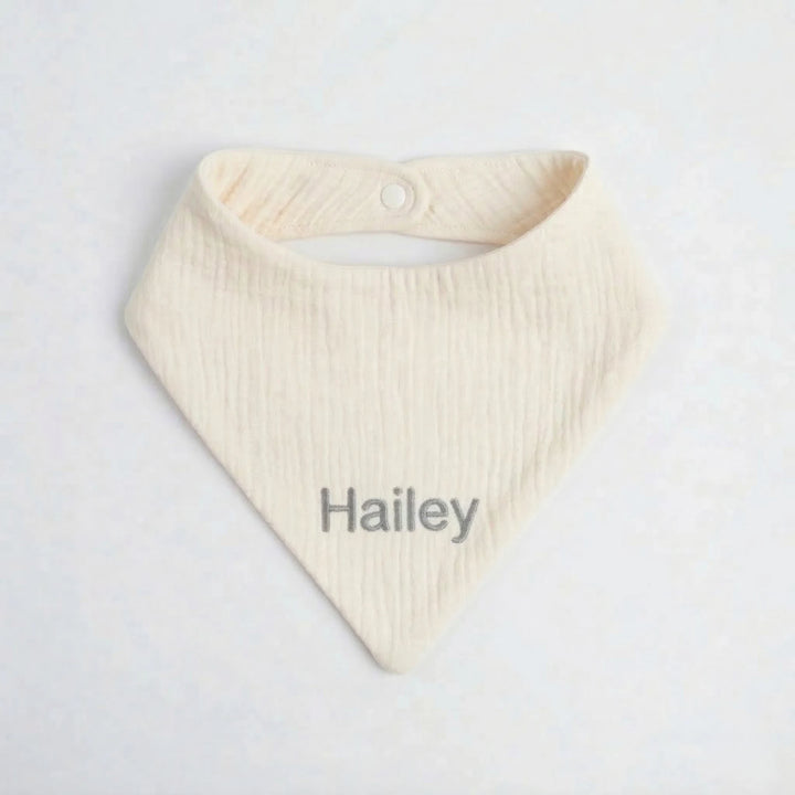 Personalised cream muslin baby bib embroidered with the name “Hailey” in grey thread, photographed on a clean white background.
