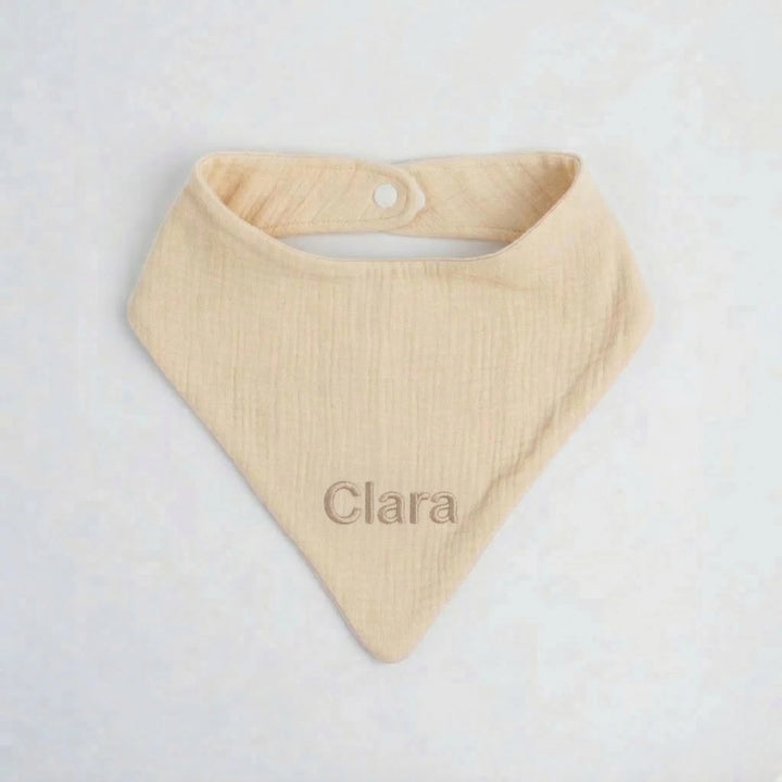 Personalised light beige muslin baby bib embroidered with the name “Clara” in beige thread, photographed on a clean white background.