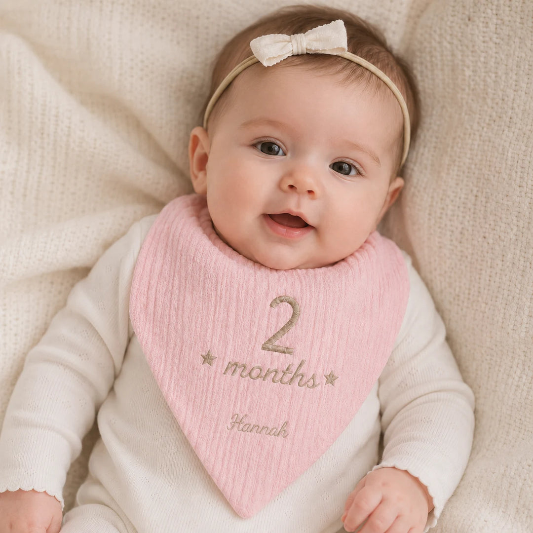 Baby wearing a pink bib with '2 months' and 'Hannah' text, lying on a beige blanket.