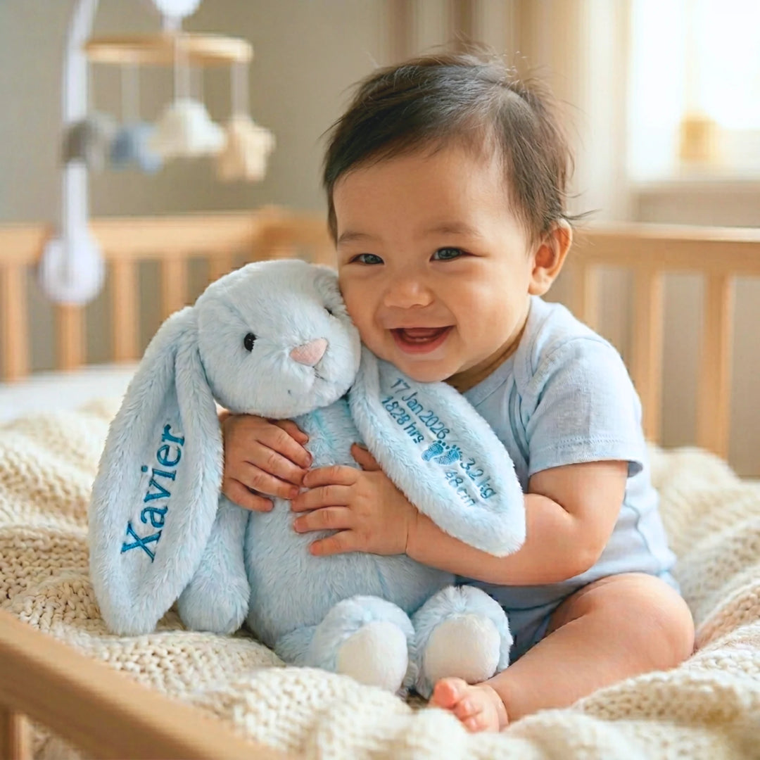 Baby holding a blue bunny toy with 'Xavier' embroidered on it, sitting in a crib.