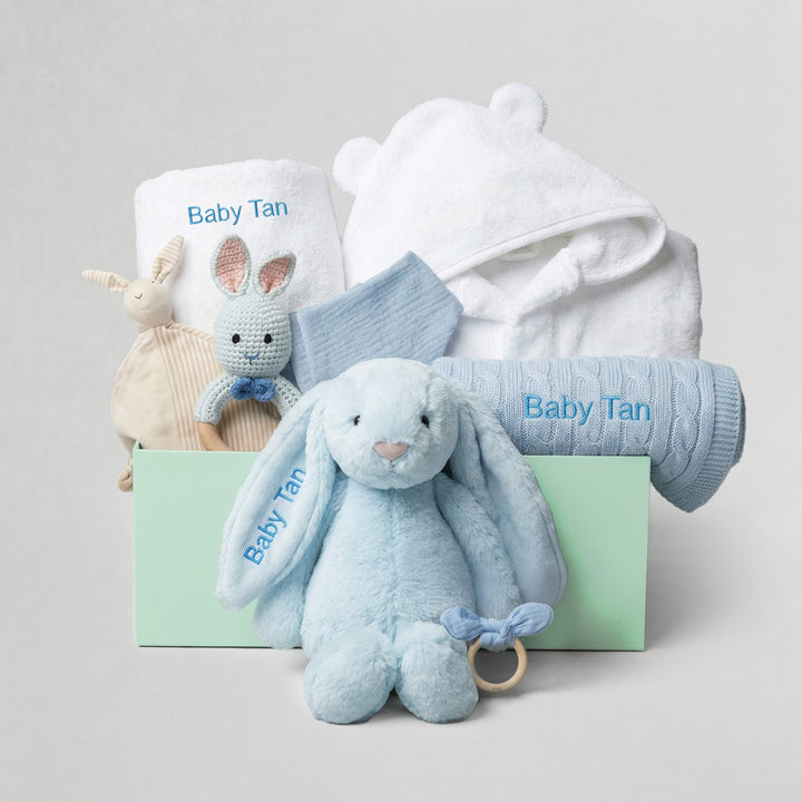 Baby gift set with blue plush bunny, white hooded towel, and soft toys on a light gray background.