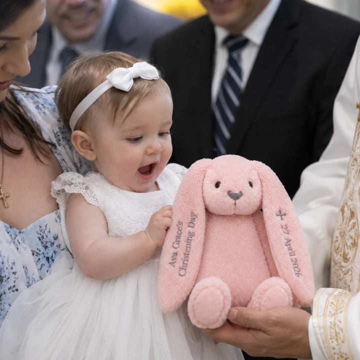 Baby holding a pink stuffed bunny with embroidered text, surrounded by adults in formal attire.