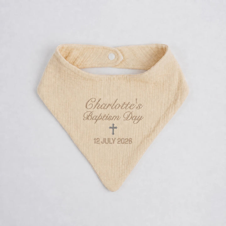 A personalized baby bandana bib in a soft cream cotton gauze fabric, featuring custom embroidery that reads 'Charlotte's Baptism Day' and '12 JULY 2026' with a small cross, laid flat on a light gray background.