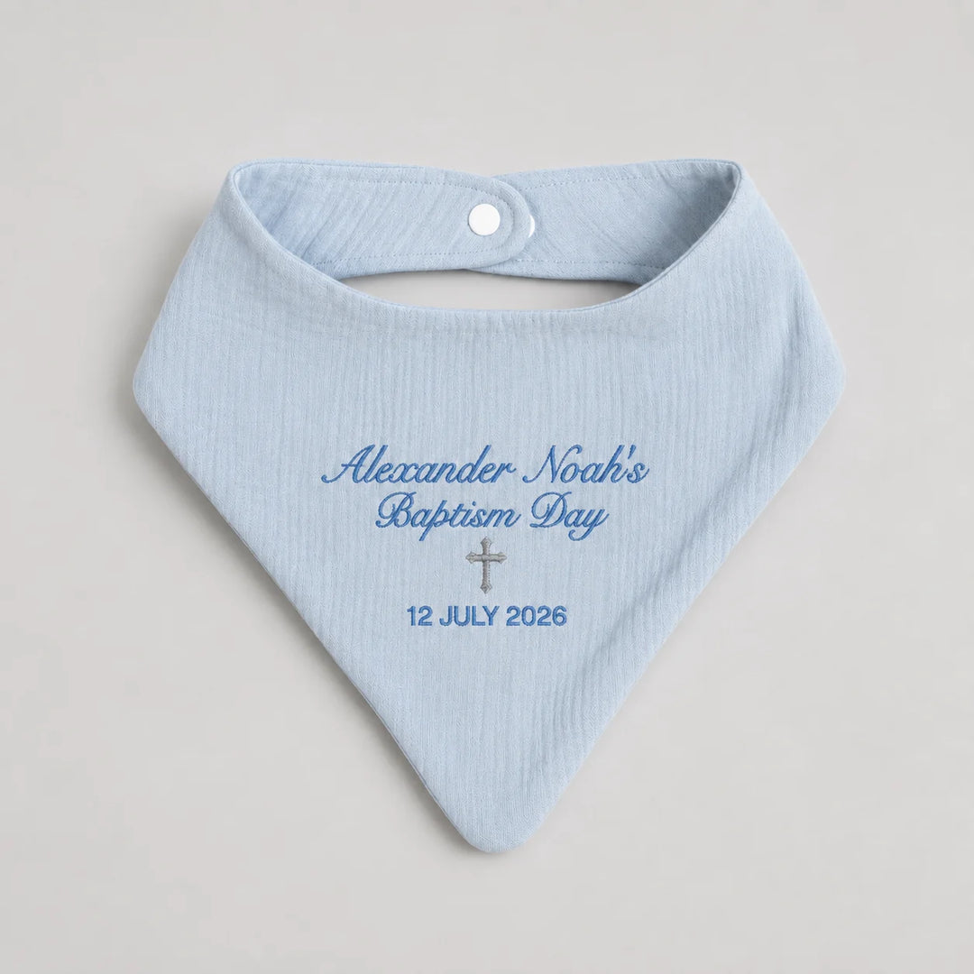 A light blue baby bandana bib with personalized blue embroidery reading 'Alexander-Noah's Baptism Day' and a small silver cross, on a neutral light gray background.