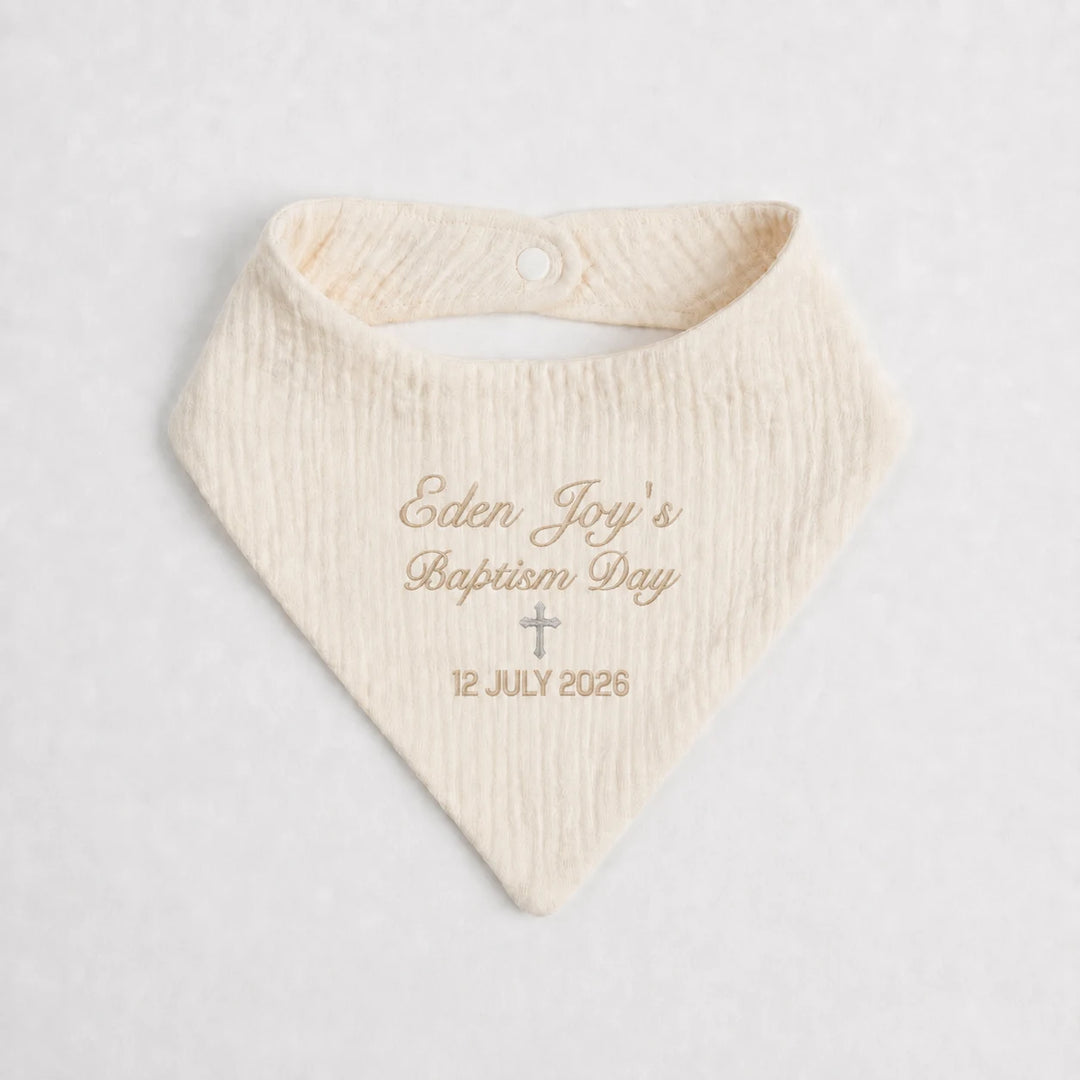 A cream-colored textured baby bandana bib with personalized gold embroidery reading 'Eden Joy's Baptism Day' and the date '12 JULY 2026' with a small cross, displayed on a light gray background.