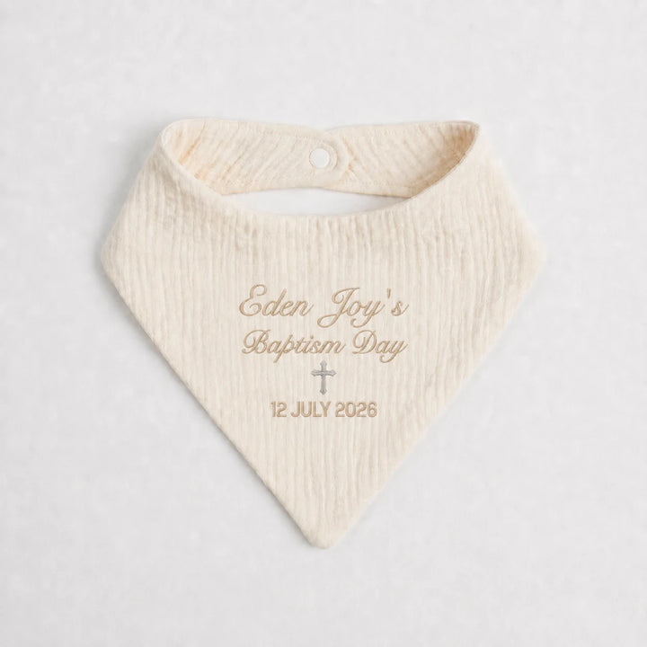 A cream-colored textured baby bandana bib with personalized gold embroidery reading 'Eden Joy's Baptism Day' and the date '12 JULY 2026' with a small cross, displayed on a light gray background.