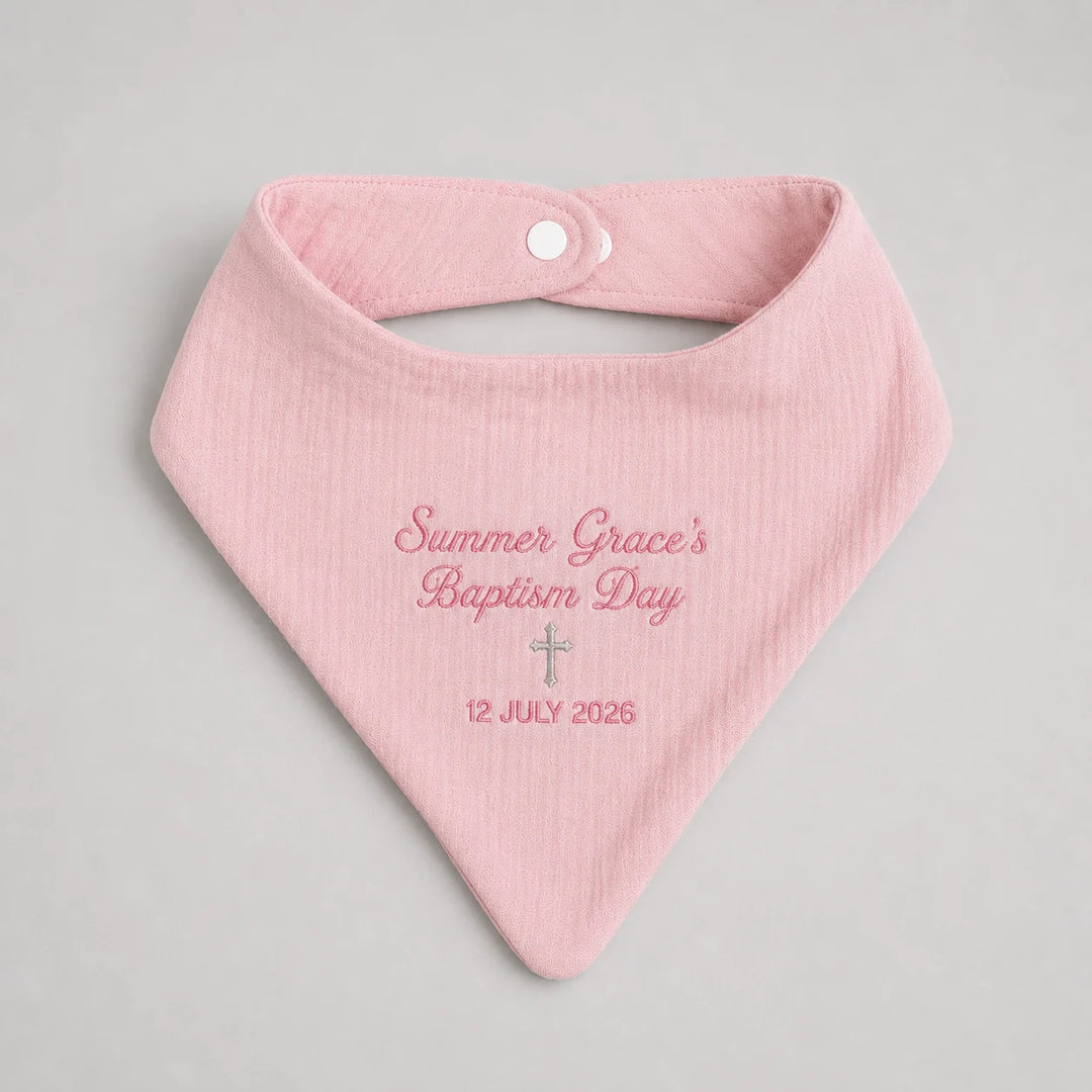 A light pink muslin baby bib with custom embroidered text reading 'Summer Grace's Baptism Day' and a date '12 JULY 2026' on a neutral gray background.