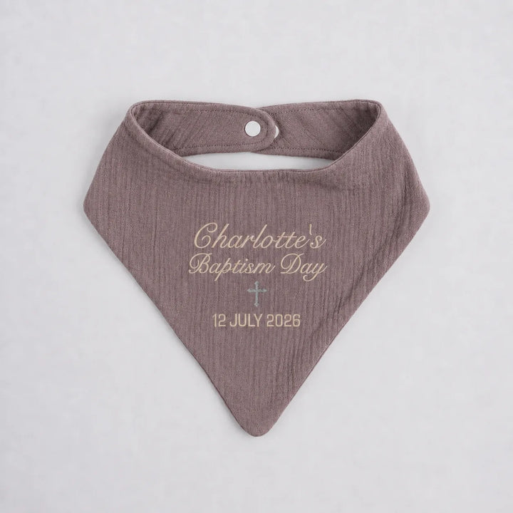 A personalized muslin cotton bandana bib in a dusty mauve color, featuring custom embroidery that reads 'Charlotte's Baptism Day' and the date '12 JULY 2025' with a small cross, shown on a light gray background.
