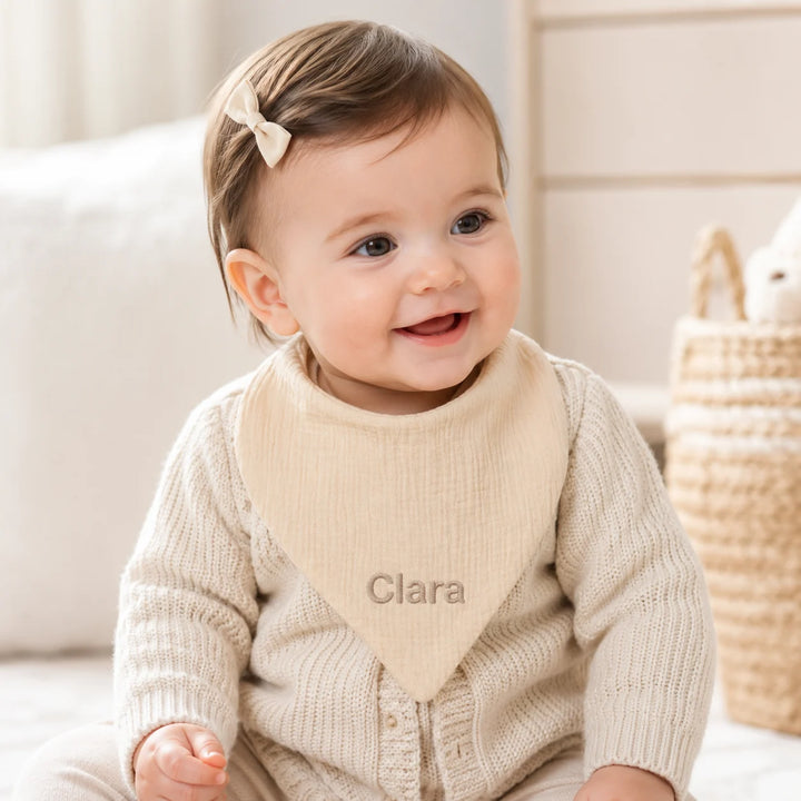 Smiling baby girl wearing a personalised light beige muslin bib embroidered with the name “Clara,” photographed in a softly lit neutral nursery.