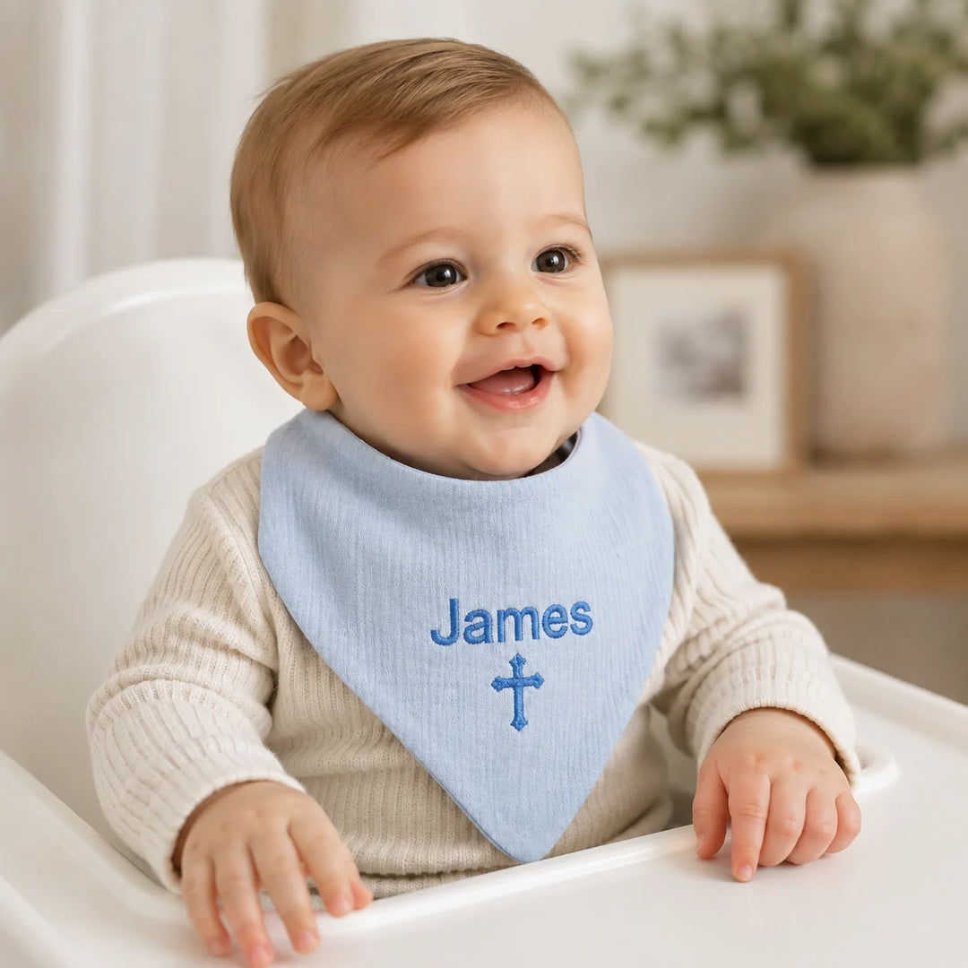 A smiling baby sitting in a white high chair wearing a light blue bib with the name 'James' and a cross embroidered on the front.