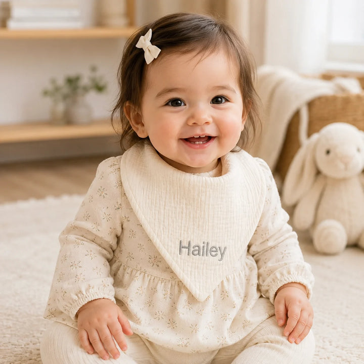 Smiling baby girl wearing a personalised cream muslin bib embroidered with the name “Hailey” in grey thread, photographed in a warm, softly lit neutral nursery setting.
