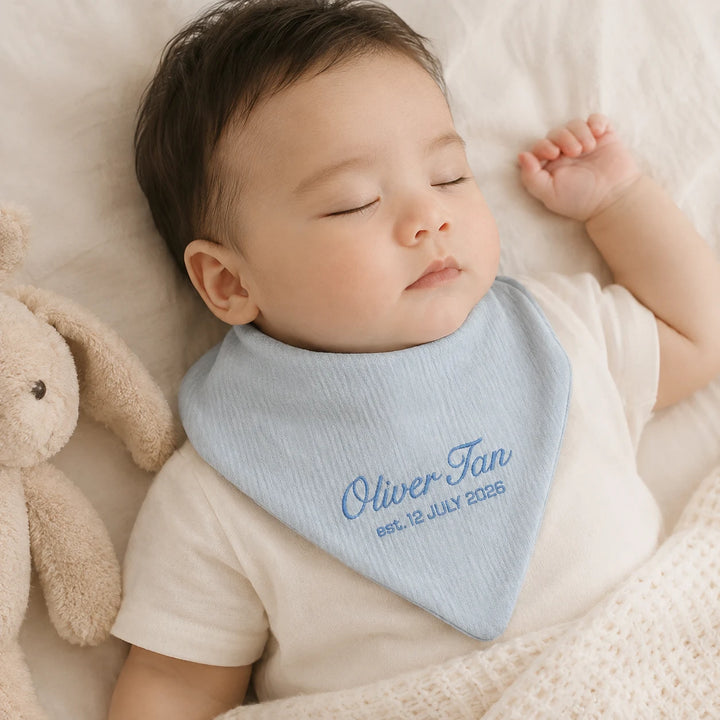 A sleeping baby wearing a light blue personalised bib with the name 'Oliver Tan' and the date 'est. 12 July 2025' embroidered in blue, lying on a soft cream blanket next to a plush bunny toy.