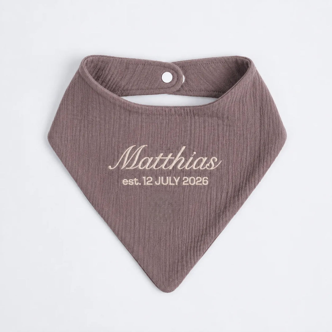 A personalized baby bandana bib in a textured plum color, featuring the name 'Matthias' and the date 'est. 12 JULY 2026' printed in a light beige font on the front.