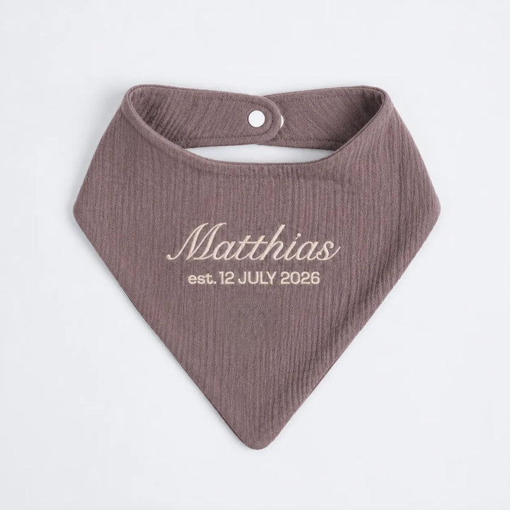 A personalized baby bandana bib in a textured plum color, featuring the name 'Matthias' and the date 'est. 12 JULY 2026' printed in a light beige font on the front.