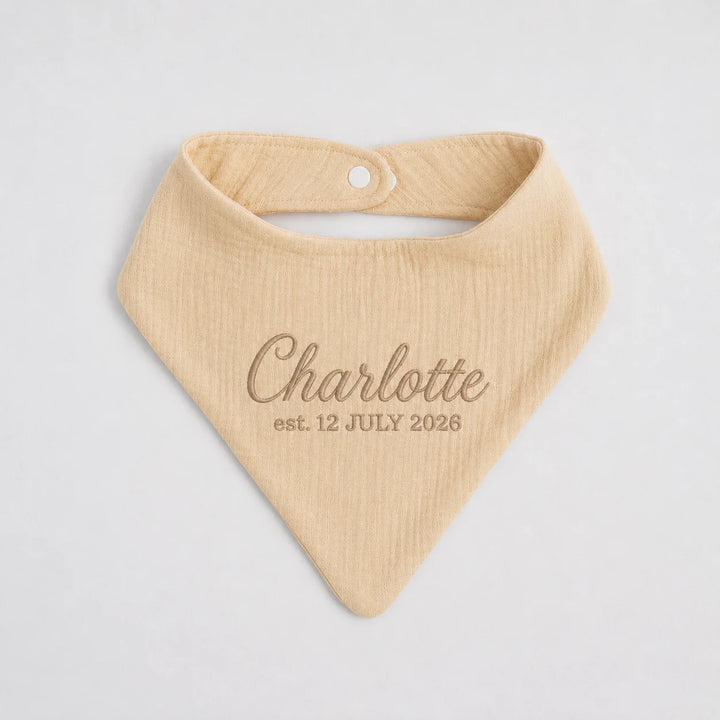 A personalized baby bib in a soft, textured cream-beige cotton gauze, featuring the name 'Charlotte' and the date 'est. 12 JULY 2026' embroidered in a matching brown tone on the front.