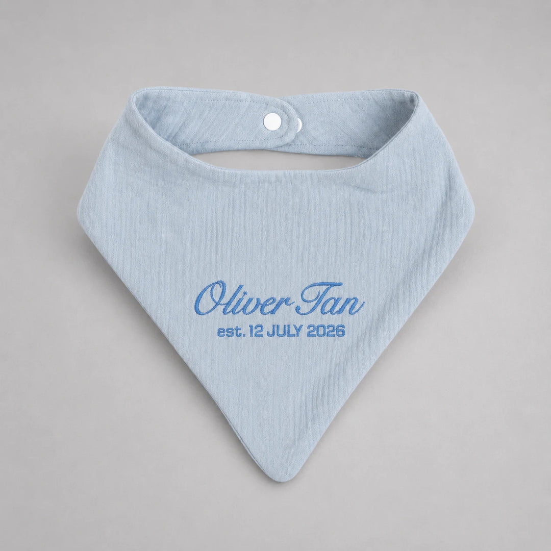 A light blue ribbed cotton bandana bib with a white snap button closure, personalized with the text 'Oliver Tan est. 12 JULY 2026' in blue embroidery, shown on a neutral gray background.