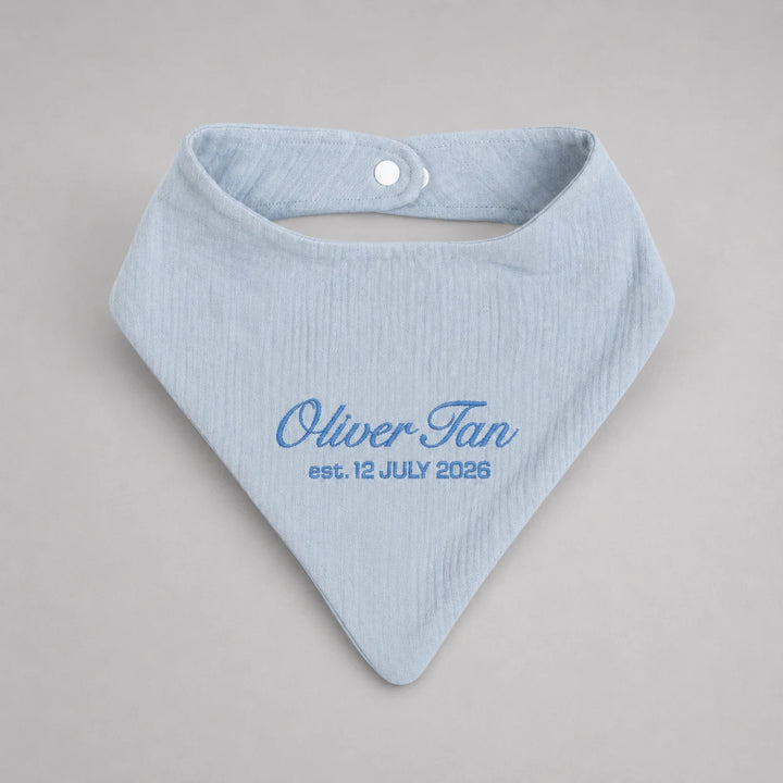 A light blue ribbed cotton bandana bib with a white snap button closure, personalized with the text 'Oliver Tan est. 12 JULY 2026' in blue embroidery, shown on a neutral gray background.