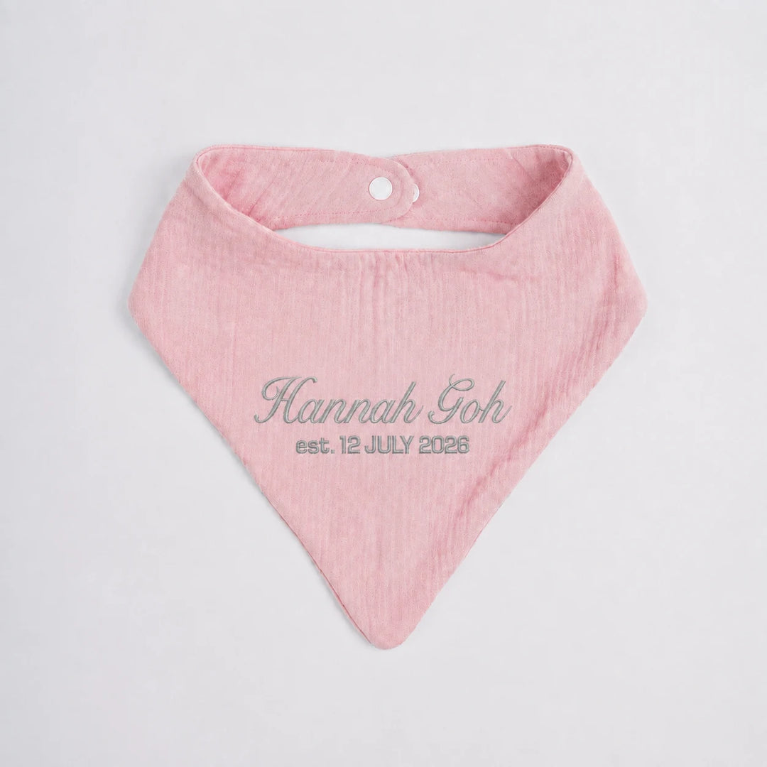 A light pink baby bandana bib with custom grey embroidered text reading 'Hannah Goh est. 12 JULY 2026' on a clean white background.