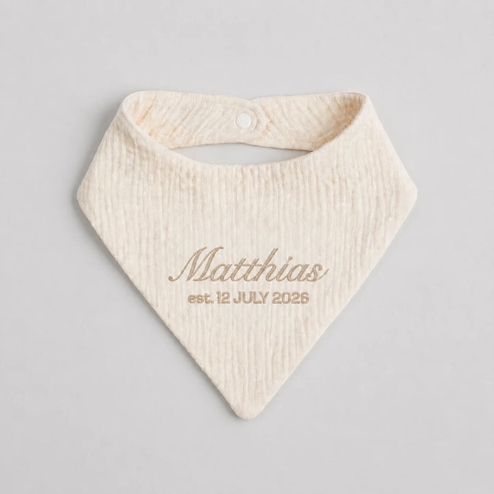 A personalized cream-colored cotton bandana bib with a textured crinkle fabric, featuring the name 'Matthias' and the date 'est. 12 JULY 2026' printed in a matching beige script on a light gray background.