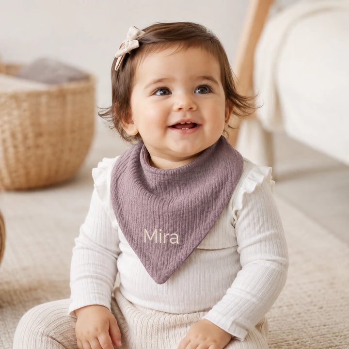 A baby wearing a personalized dusty purple ribbed bib with the name 'Mira' embroidered in cream, sitting in a softly lit nursery.