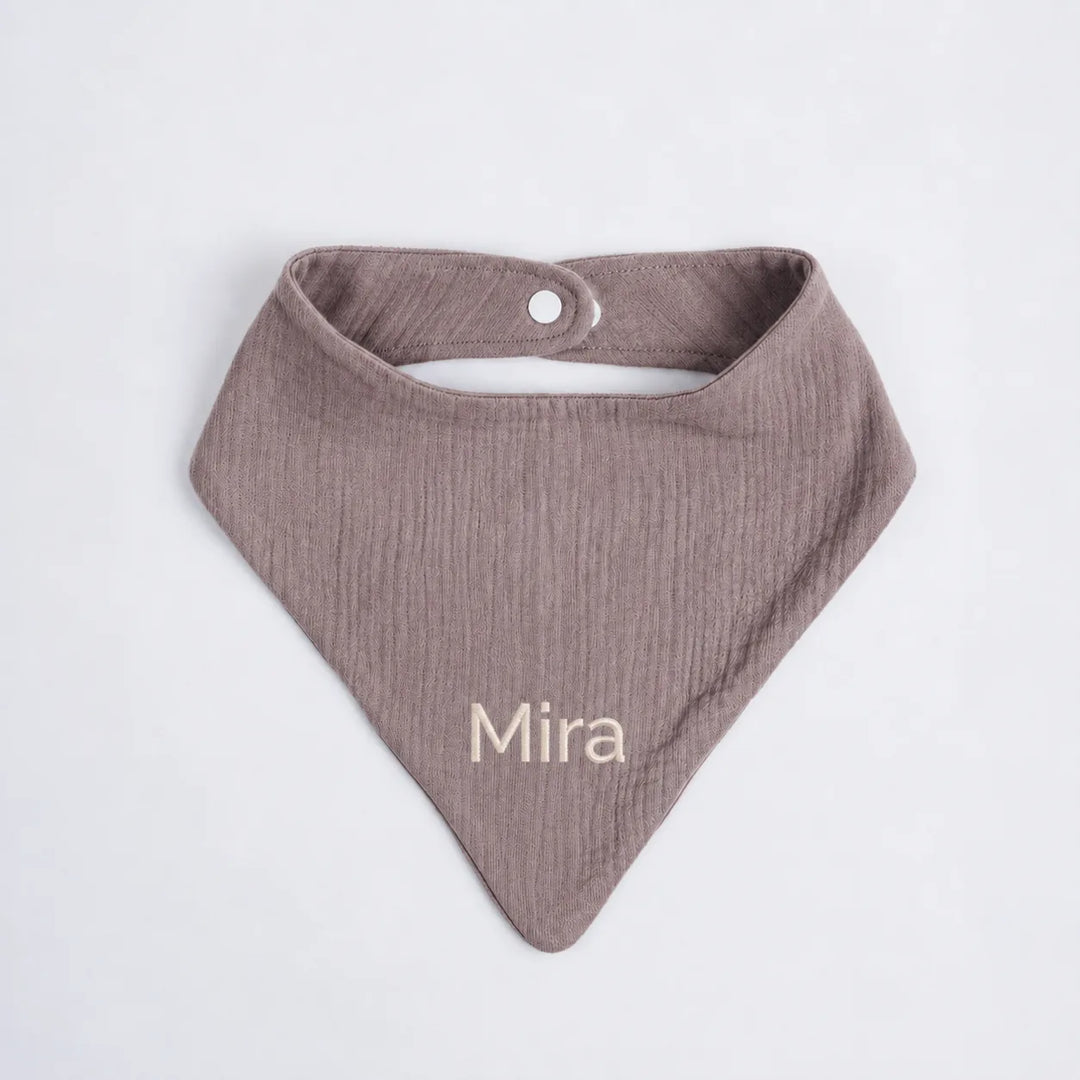 A dusty mauve cotton muslin baby bib with the name 'Mira' embroidered in cream on the front, displayed flat against a light gray background.