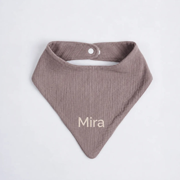 A dusty mauve cotton muslin baby bib with the name 'Mira' embroidered in cream on the front, displayed flat against a light gray background.