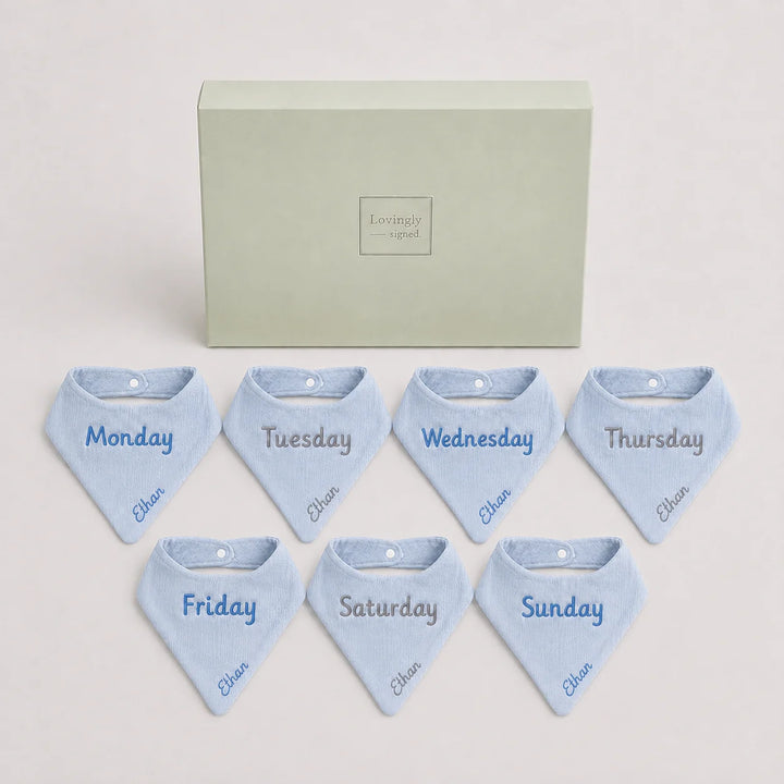Set of seven blue baby bibs with days of the week embroidery on a light gray background