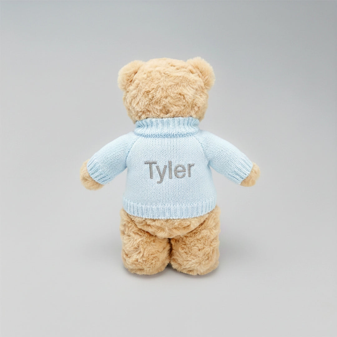 Teddy bear wearing a light blue sweater with 'Tyler' embroidered on it against a gray background