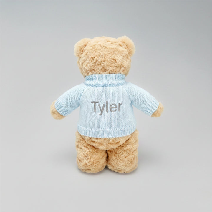 Teddy bear wearing a light blue sweater with 'Tyler' embroidered on it against a gray background