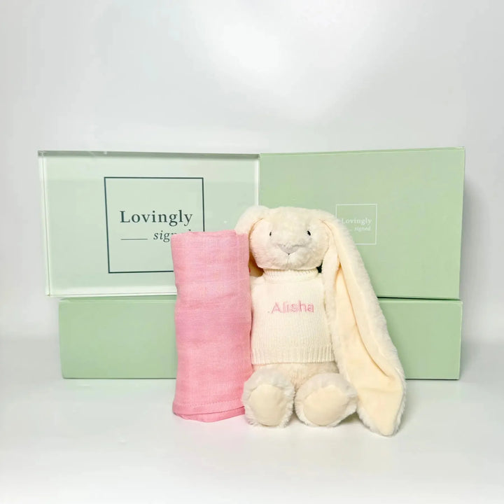 Pink swaddle blanket and cream bunny toy with 'Alisha' embroidery in front of a green gift box.
