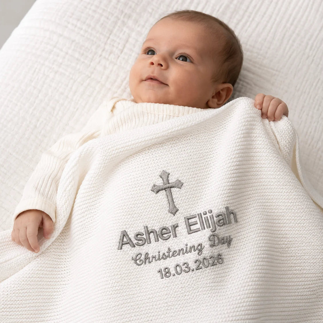 Baby wrapped in a white blanket with embroidered text on a neutral background