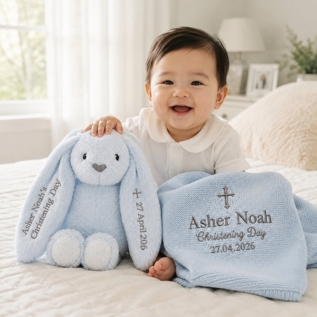 A smiling baby in a white outfit sits on a bed holding a light blue plush bunny and a matching knitted blanket. The items are personalized with the name 'Asher Noah', 'Christening Day', and the date '27.