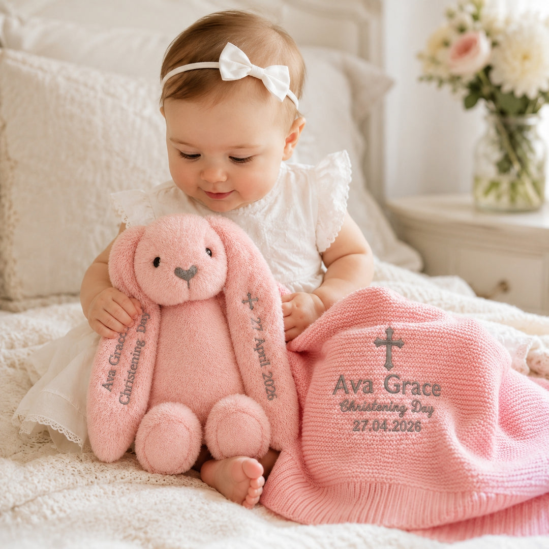 A baby girl in a white dress and bow headband sits on a bed holding a pink plush bunny, with a matching pink knitted hat placed beside her. The hat is embroidered with a cross and the text 'Ava Grace Christening Day 27.