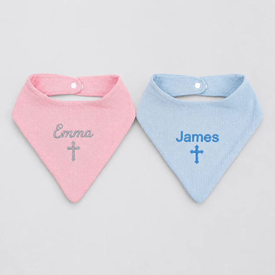 Two personalized baby bandana bibs, one light pink with the name 'Emma' and a silver cross, and one light blue with the name 'James' and a blue cross, displayed side-by-side on a light gray background.