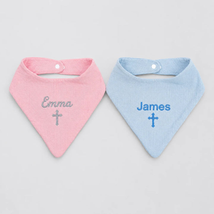 Two personalized baby bandana bibs, one light pink with the name 'Emma' and a silver cross, and one light blue with the name 'James' and a blue cross, displayed side-by-side on a light gray background.