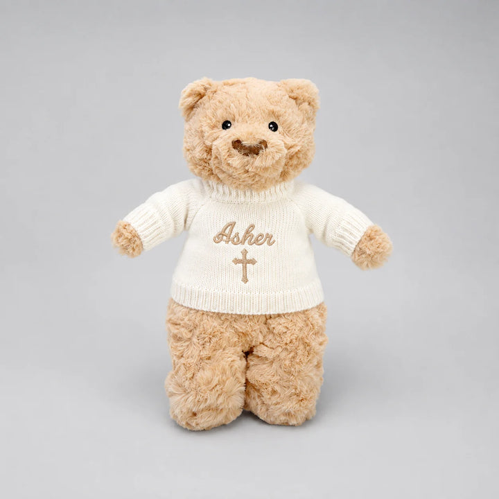 A plush teddy bear wearing a cream-colored knit sweater with the name 'Asher' and a cross embroidered on the front, set against a solid gray background.