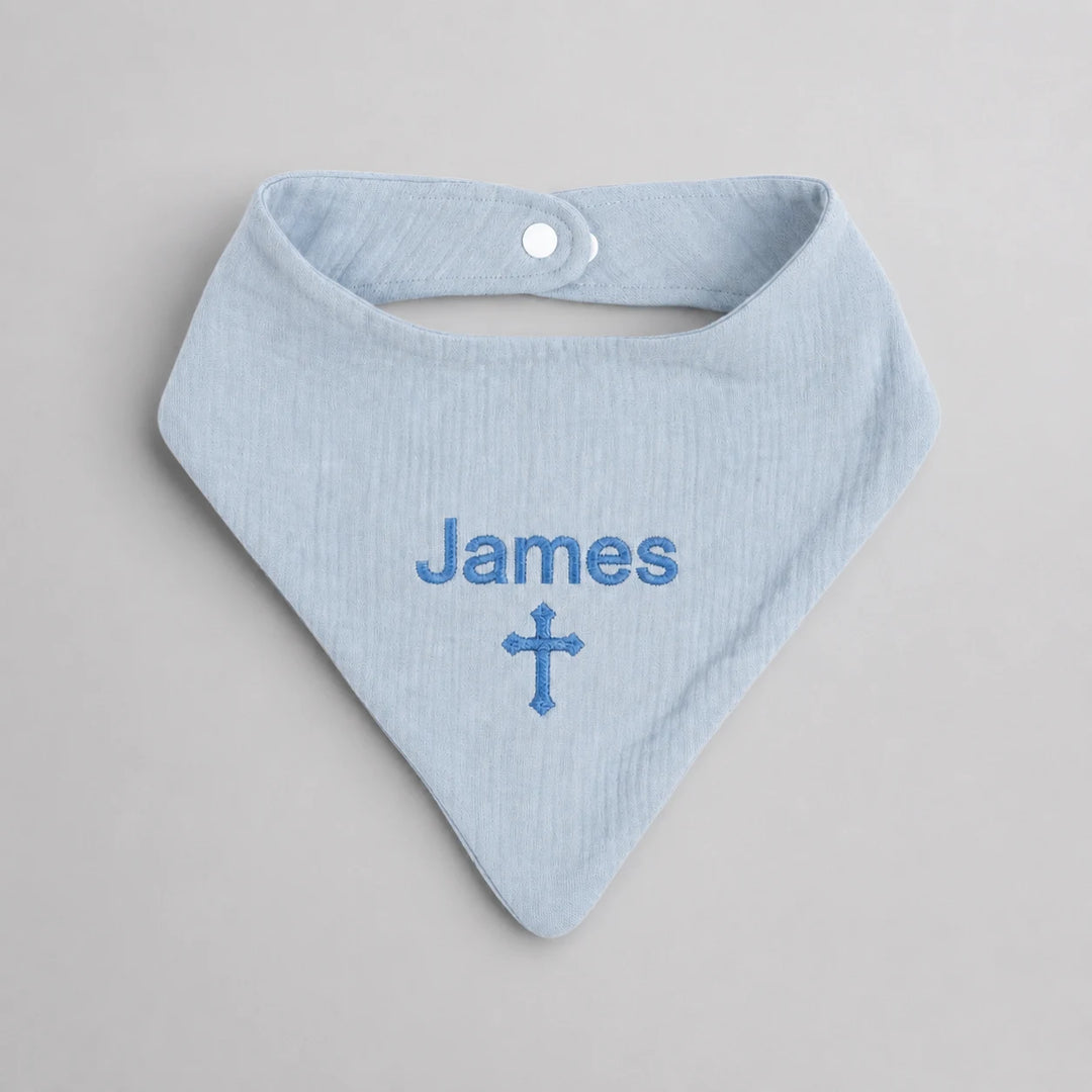 A light blue baby bandana bib with 'James' and a cross embroidered in blue on the front, displayed flat on a neutral gray background.
