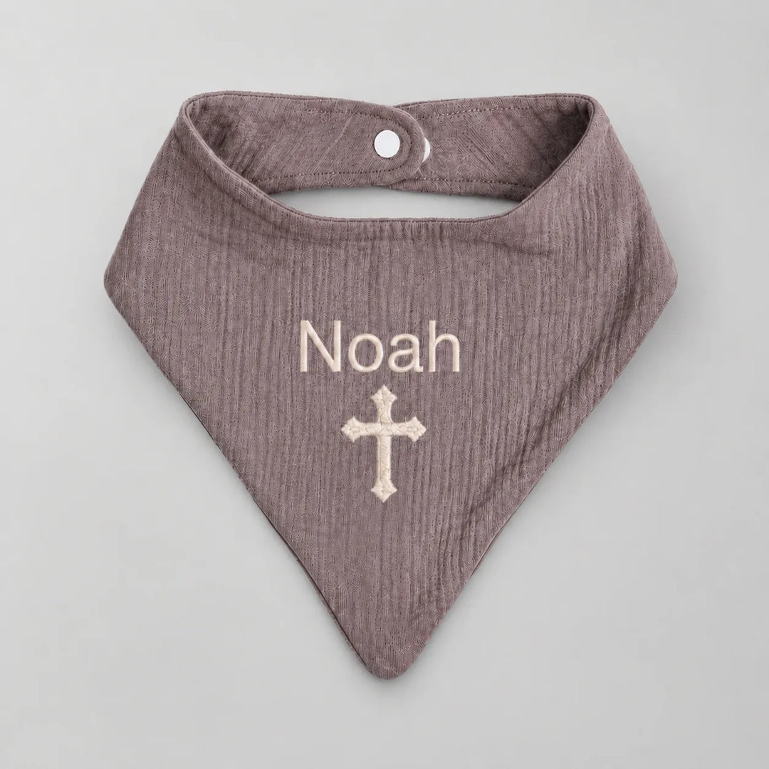 A personalized baby bandana bib in a textured dusty mauve color, featuring the name 'Noah' and a cross embroidered in cream on the front, displayed on a neutral gray background.