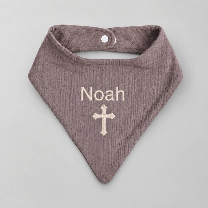 A personalized baby bandana bib in a textured dusty mauve color, featuring the name 'Noah' and a cross embroidered in cream on the front, displayed on a neutral gray background.