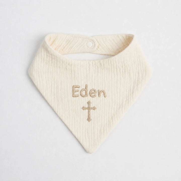 An off-white muslin baby bib with the name 'Eden' and a small cross embroidered in a light tan thread, displayed on a clean white background.