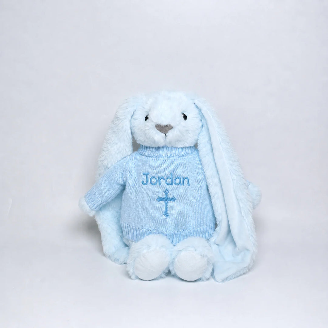 A light blue plush bunny doll wearing a matching light blue knitted sweater with the name 'Jordan' and a cross embroidered on the front, photographed against a plain light gray background.