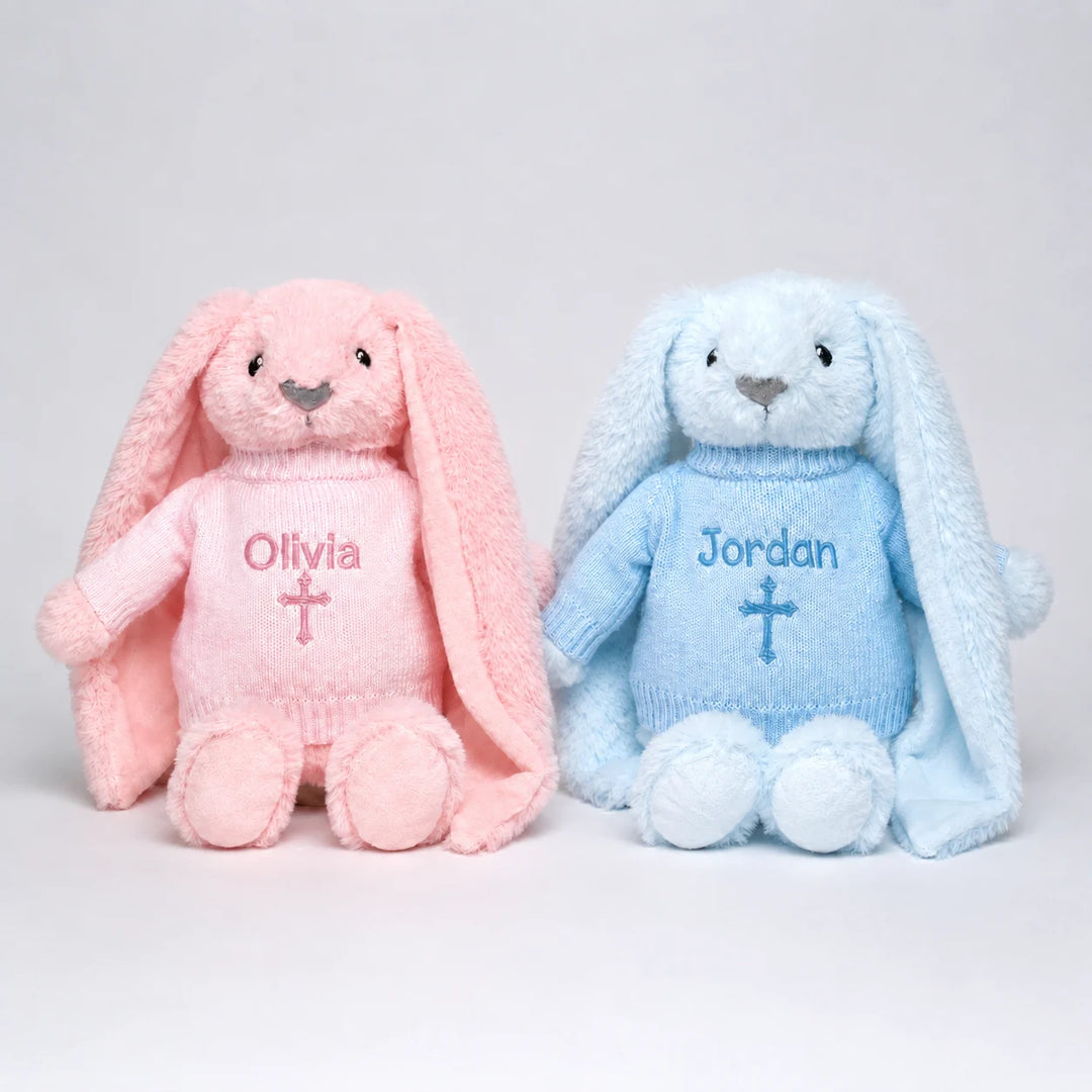 Two plush bunny toys side by side, one pink and one light blue, each wearing a knitted sweater with a cross and personalized name (Olivia and Jordan).